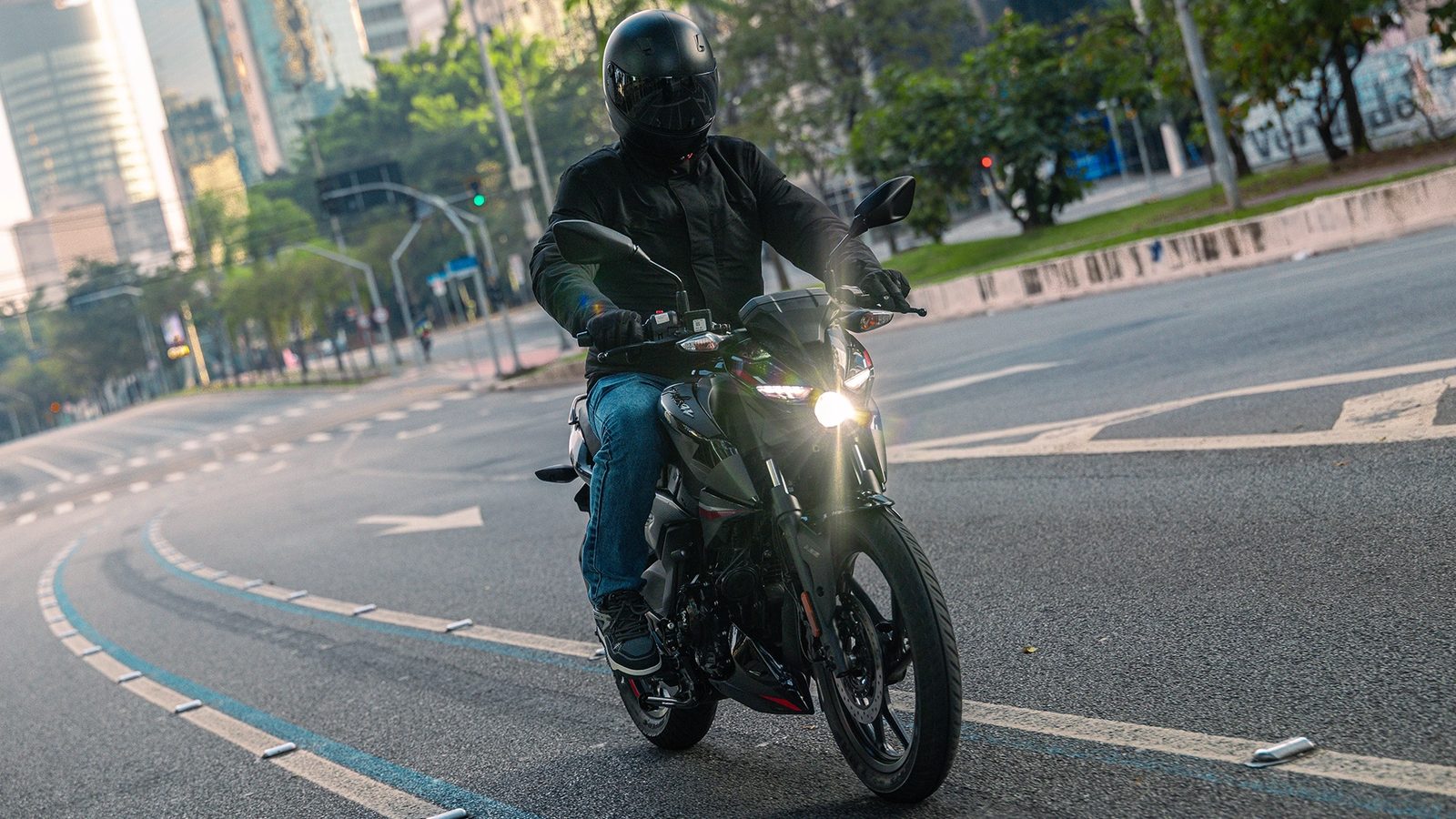 Piloto na Bajaj Pulsar N150 preta, com farol aceso, em uma avenida urbana